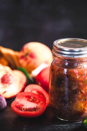 Jar of roasted peach salsa with fresh peaches, tomatoes, red onion, garlic, and jalapeño on a dark surface.