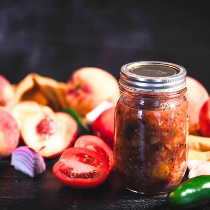 Jar of roasted peach salsa with fresh peaches, tomatoes, red onion, garlic, and jalapeño on a dark surface.