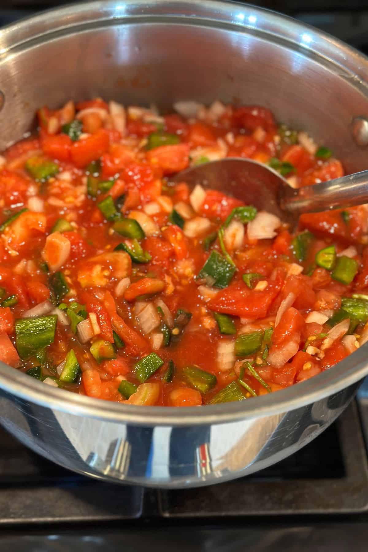 Homemade salsa simmering on the stovetop with chopped tomatoes, onions, and green peppers in a stainless steel pot