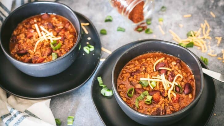 Two bowls of venison chili on a table topped with cheddar cheese and green onions. 