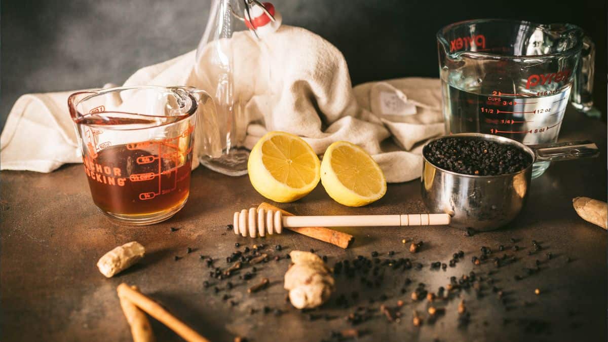 A rustic preparation scene for elderberry syrup, featuring a glass measuring cup of honey, halved lemons, a metal measuring cup filled with dried elderberries, fresh ginger, cinnamon sticks, cloves, and a honey dipper. A swing-top bottle and a second measuring cup with water sit in the background on a warm-toned surface, surrounded by scattered spices and a cream-colored cloth.