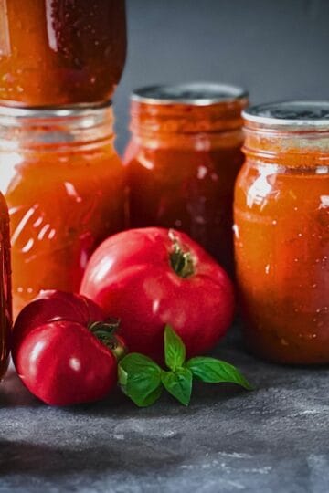 Jars of home-canned tomato soup surrounded by fresh tomatoes and basil on a dark countertop.