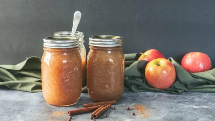 Jars of home canned applesauce on a counter surrounded by fresh apples, cinnamon sticks, allspice and nutmeg. 