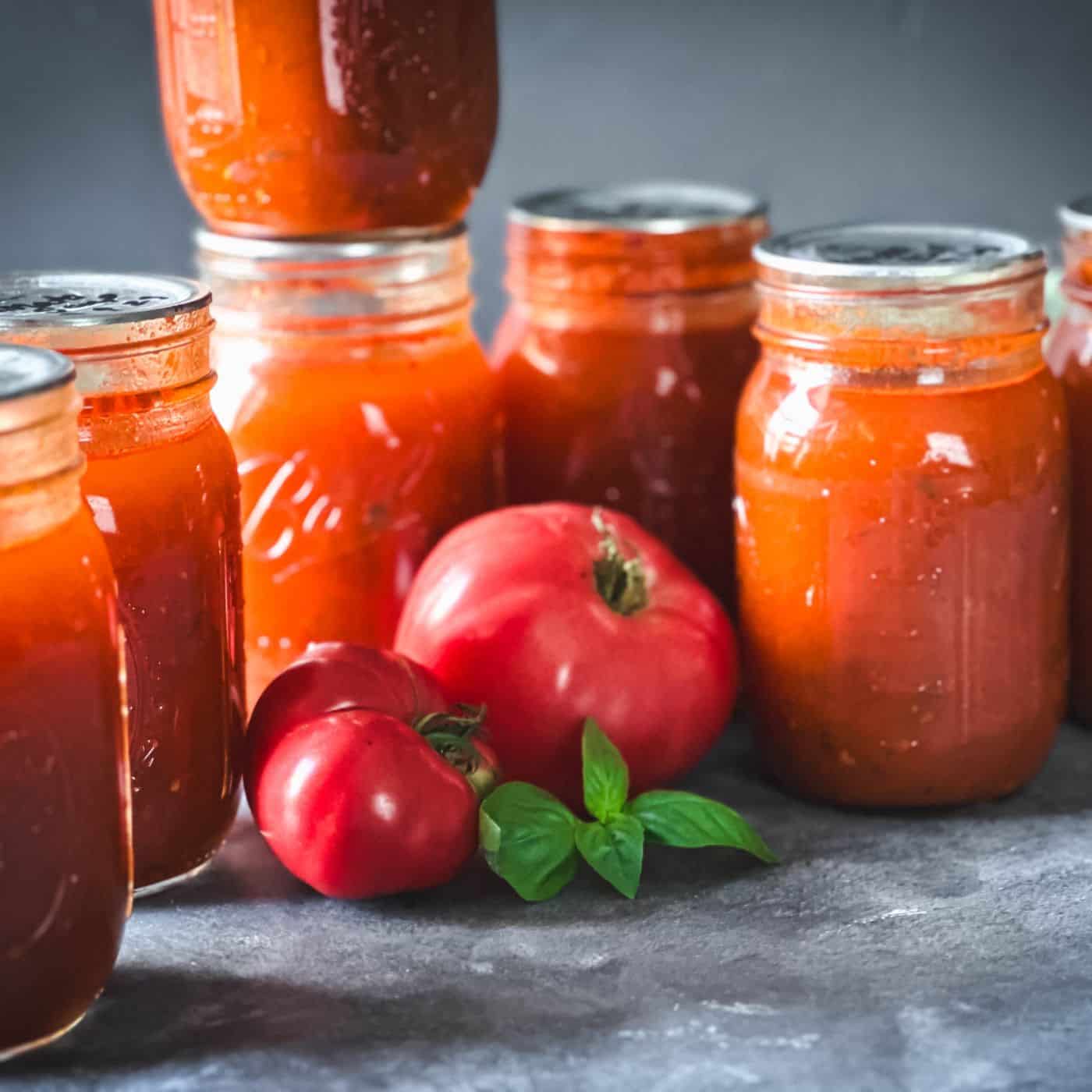 Jars of home canned tomato soup sitting on a counter with fresh tomatoes and basil. Canning tomato soup at home like this is easy.