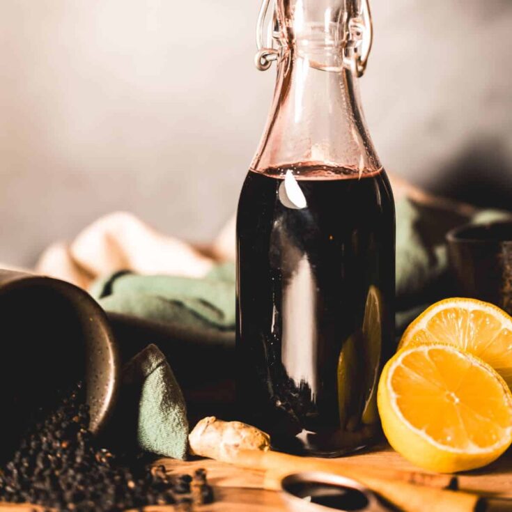 A rustic glass bottle filled with deep purple elderberry syrup, surrounded by fresh lemon halves, a piece of ginger, and a wooden spoon with elderberries scattered on a wooden surface. The scene is warmly lit, emphasizing the natural ingredients and homemade feel of the syrup.
