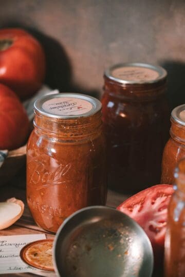 Glass mason jars filled with homemade tomato soup surrounded by fresh tomatoes, onion, a canning ladle, and a handwritten recipe.