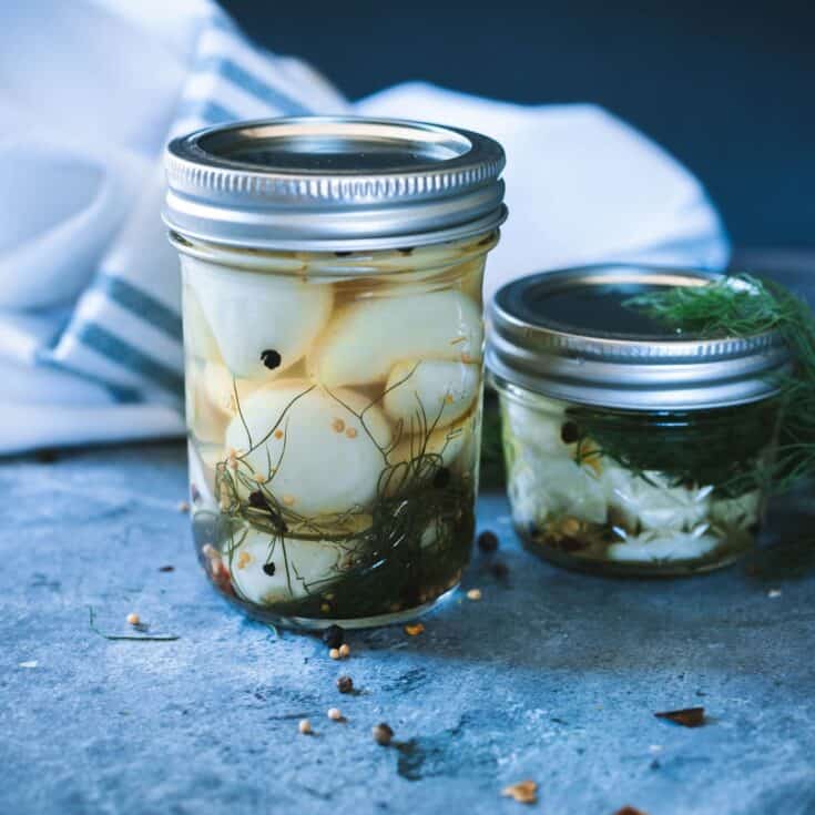 Jars of pickled garlic on a counter surrounded by spices and fresh dill.