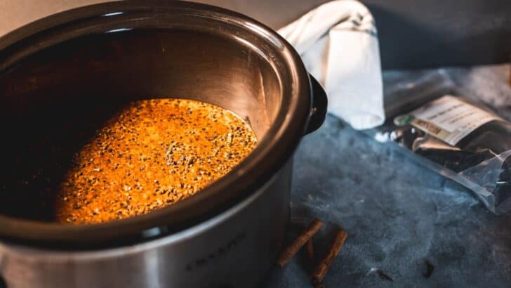 A slow cooker filled with simmering homemade elderberry syrup, with dried elderberries and cinnamon sticks nearby on a dark countertop. A white kitchen towel rests in the background.