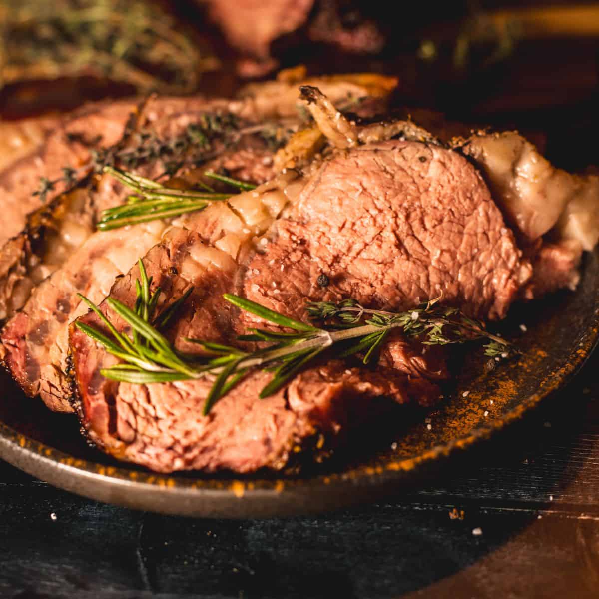 Slices of herb-crusted smoked prime rib on a rustic plate, garnished with fresh rosemary and thyme sprigs. The tender, juicy meat showcases a perfectly cooked pink center and a flavorful crust.