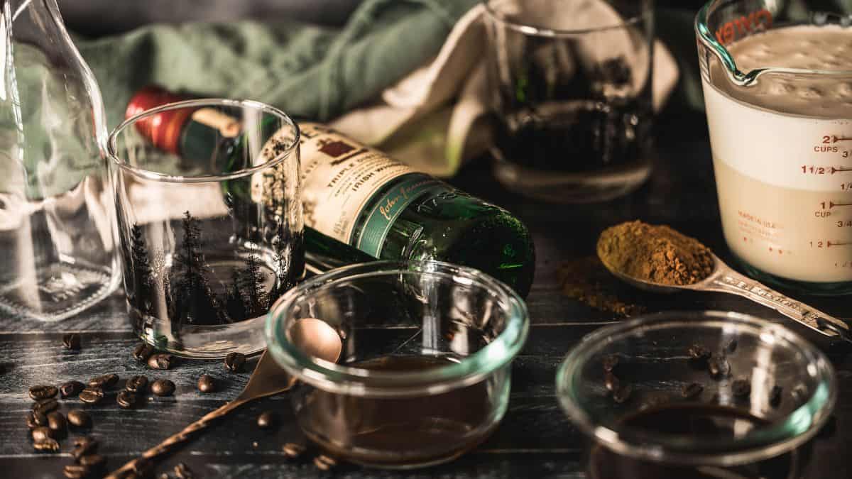 Flatlay of Irish cream ingredients including Irish whiskey, cream, chocolate syrup, coffee, and vanilla, styled on a dark wood surface with glassware and coffee beans for a rustic aesthetic.