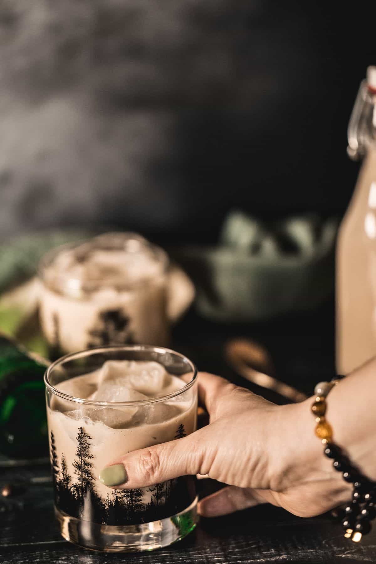 A hand holding a glass of homemade Irish cream over ice, served in a forest-print tumbler with a cozy, moody background perfect for the holidays or St. Patrick’s Day.