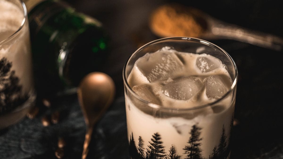 A glass of homemade Irish cream liqueur served over ice in a moody forest-print tumbler, with warm lighting and cozy, rustic background elements.