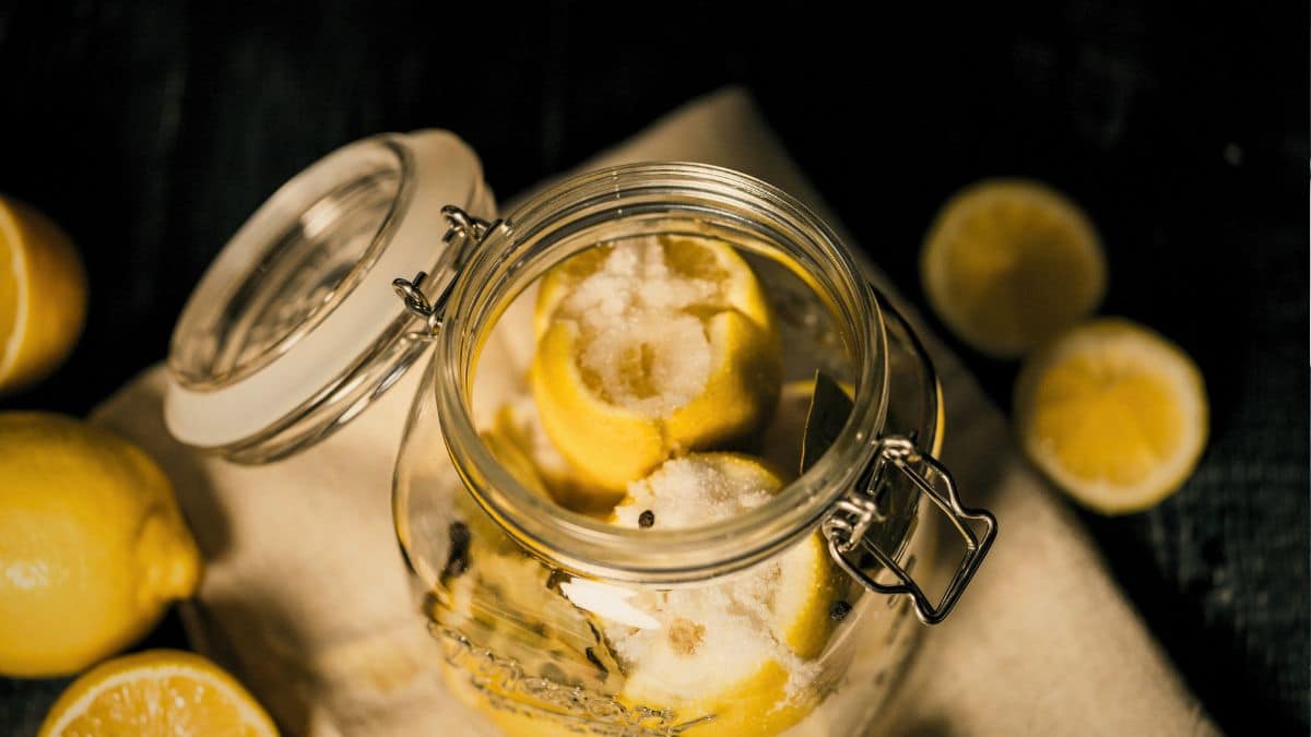 A glass jar filled with freshly salted lemons, bay leaves, and peppercorns, ready for the preserving process. The open jar sits on a rustic linen cloth with scattered lemon slices, coarse salt, and a dark moody background, capturing the early stages of making preserved lemons.