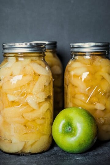Homemade canned apple slices in quart jars with fresh apples on a dark background.