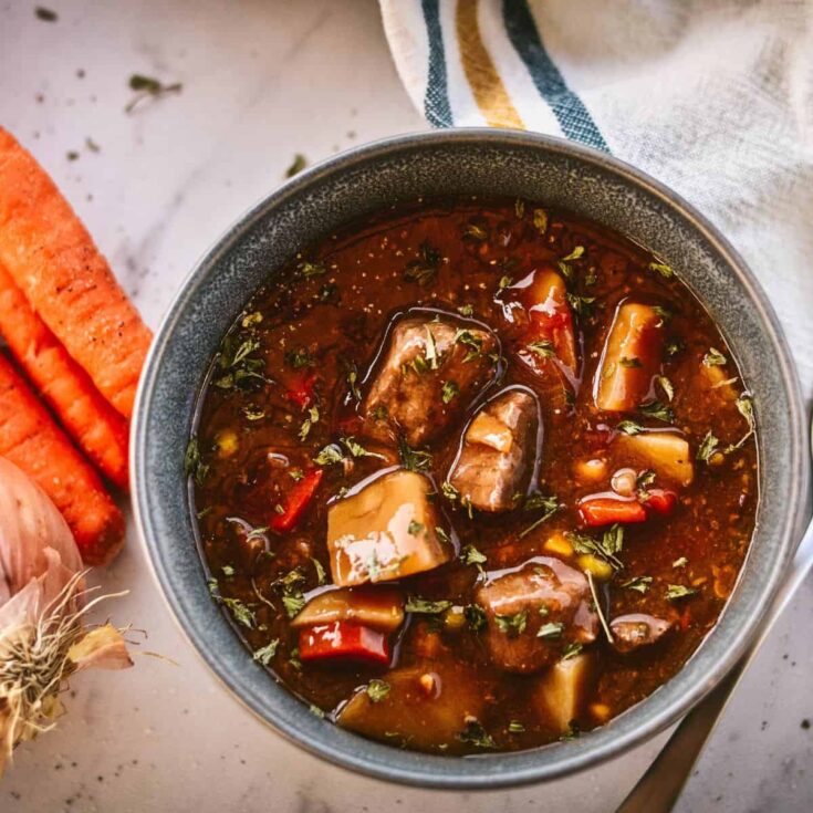 A warm and inviting bowl of hearty fall vegetable beef soup, filled with tender beef chunks, garden-fresh vegetables, and a rich broth. The soup is garnished with fresh herbs and served in a rustic bowl, with vibrant carrots and garlic resting nearby on a light-colored surface, creating a cozy and comforting autumn-inspired scene.