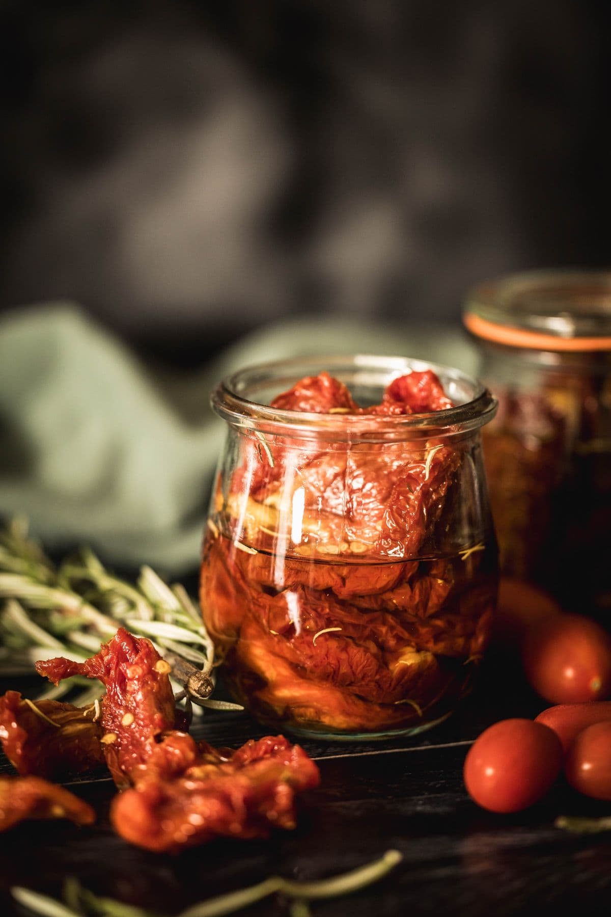 Jar of sun-dried tomatoes in olive oil with rosemary, surrounded by fresh cherry tomatoes and loose herbs on a dark wood surface.