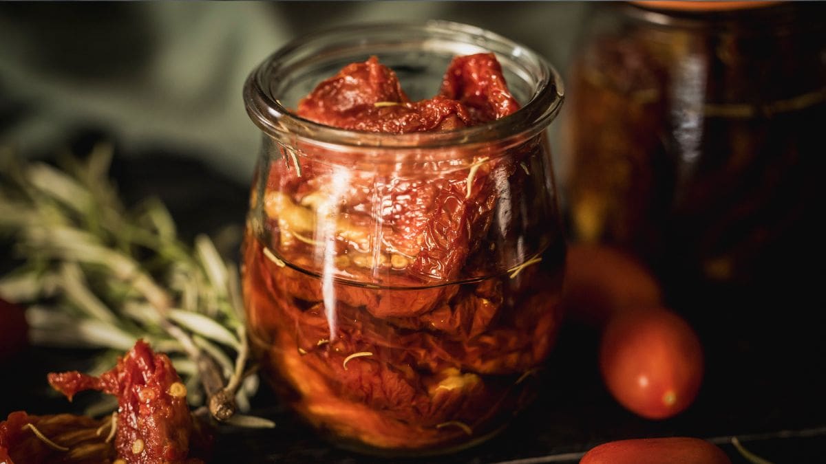 Close-up of a glass jar filled with sun-dried tomatoes in olive oil, infused with fresh rosemary, surrounded by cherry tomatoes and herbs on a dark rustic surface.