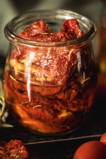 Close-up of a glass jar filled with sun-dried tomatoes in olive oil, infused with fresh rosemary, surrounded by cherry tomatoes and herbs on a dark rustic surface.