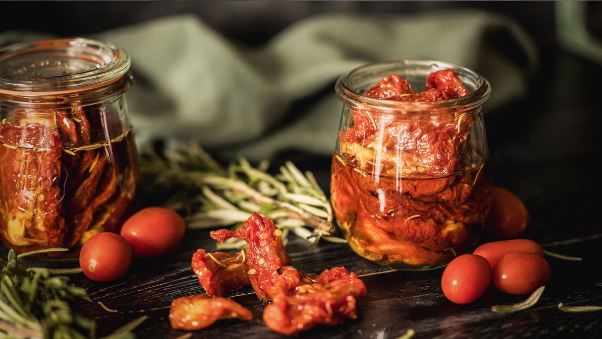 Two jars filled with sun-dried tomatoes in olive oil, surrounded by cherry tomatoes and fresh rosemary sprigs on a dark rustic table.
