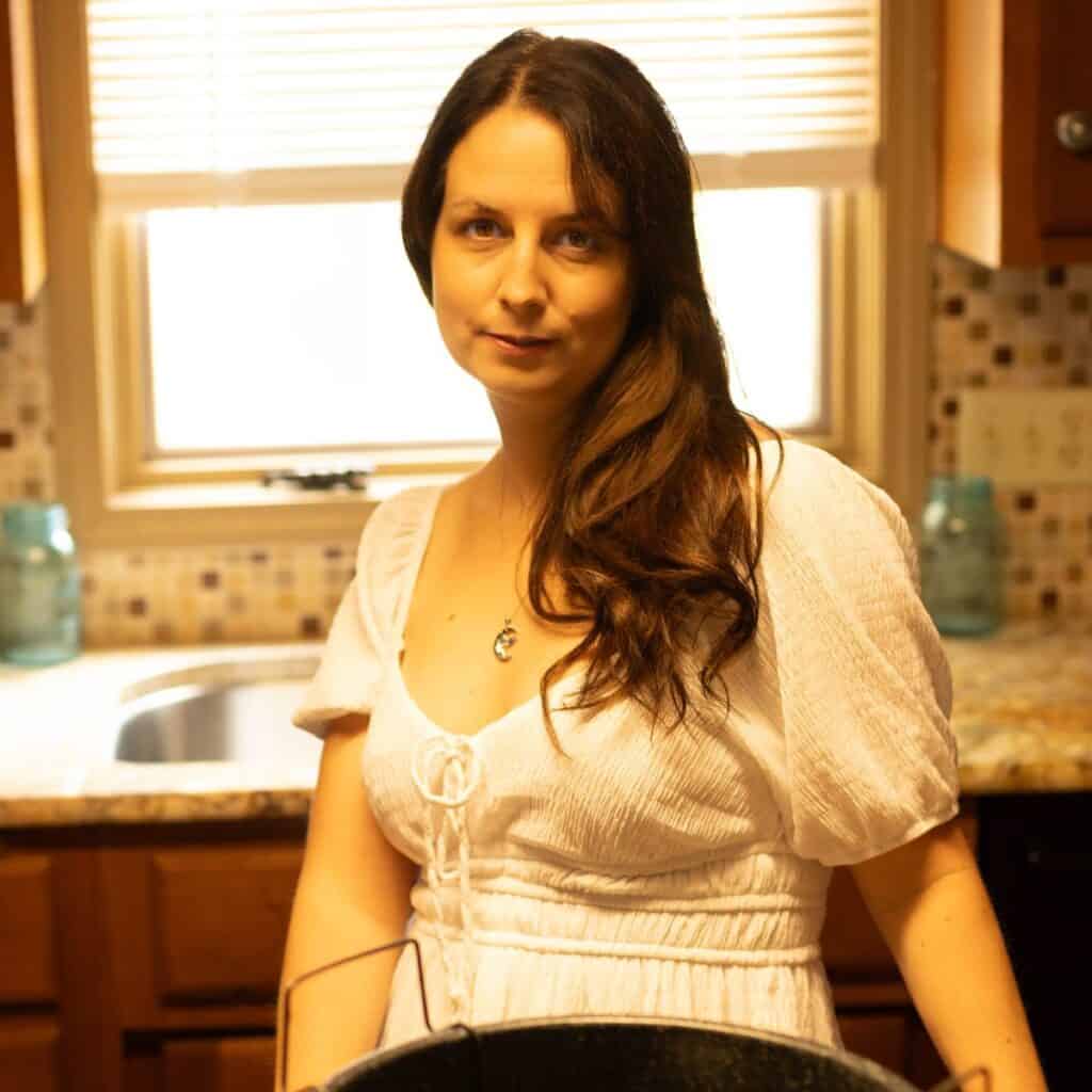 Elle McCoy stands in a warm, rustic kitchen wearing a white dress, softly smiling as sunlight streams through the window behind her. Her long brown hair cascades over one shoulder, and a canning pot and mason jars sit in the foreground.