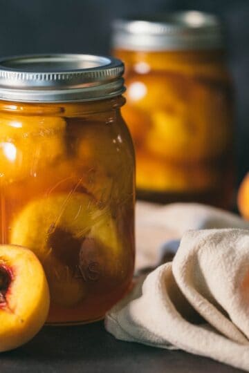 Glass mason jars filled with canned peach sweet tea surrounded by fresh peaches and a linen cloth