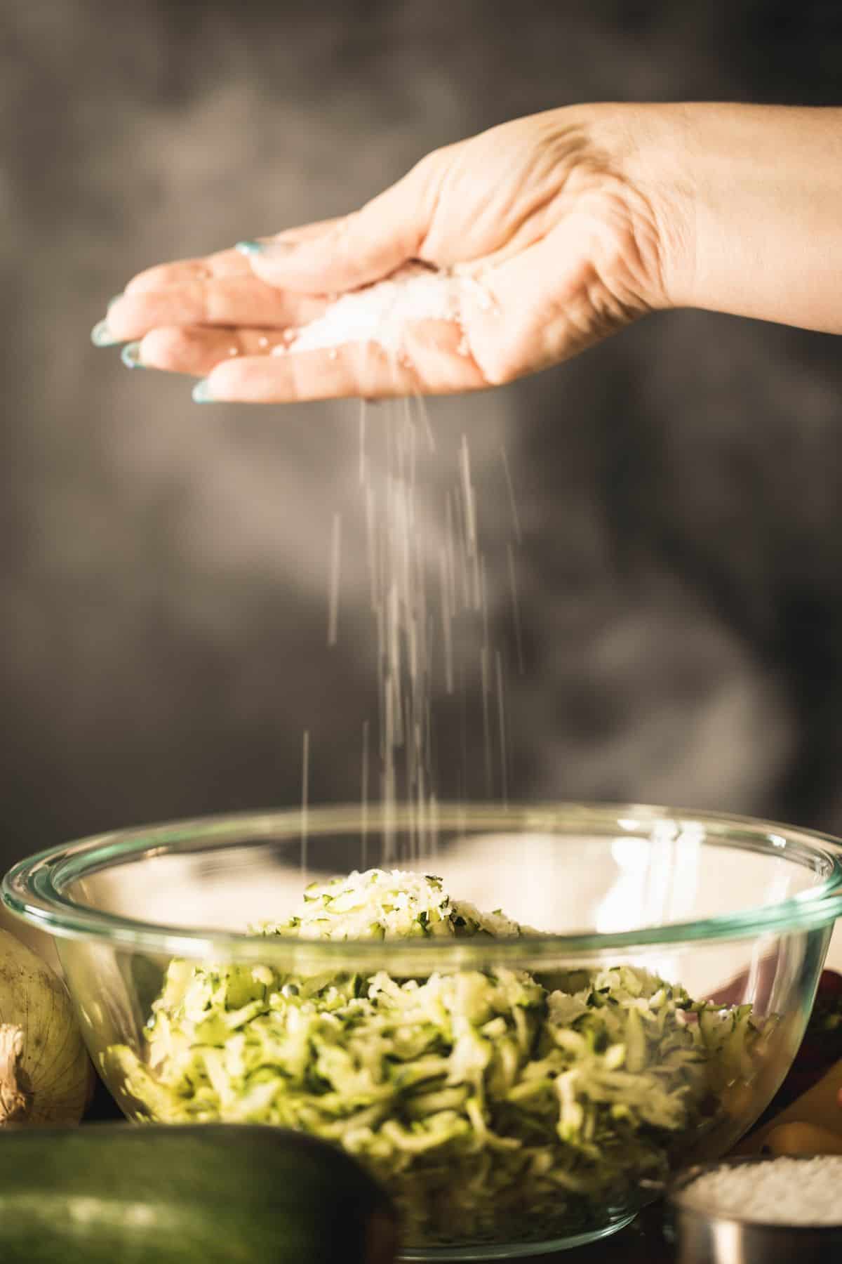 Hand sprinkling salt over a bowl of shredded zucchini