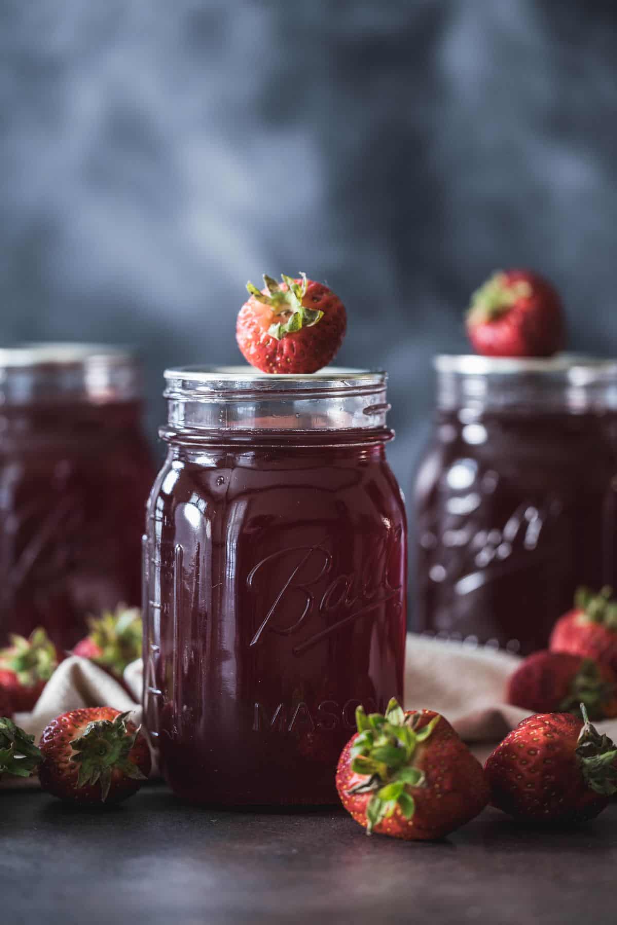 Jar of homemade canned strawberry syrup topped with a fresh strawberry, surrounded by berries on a dark background