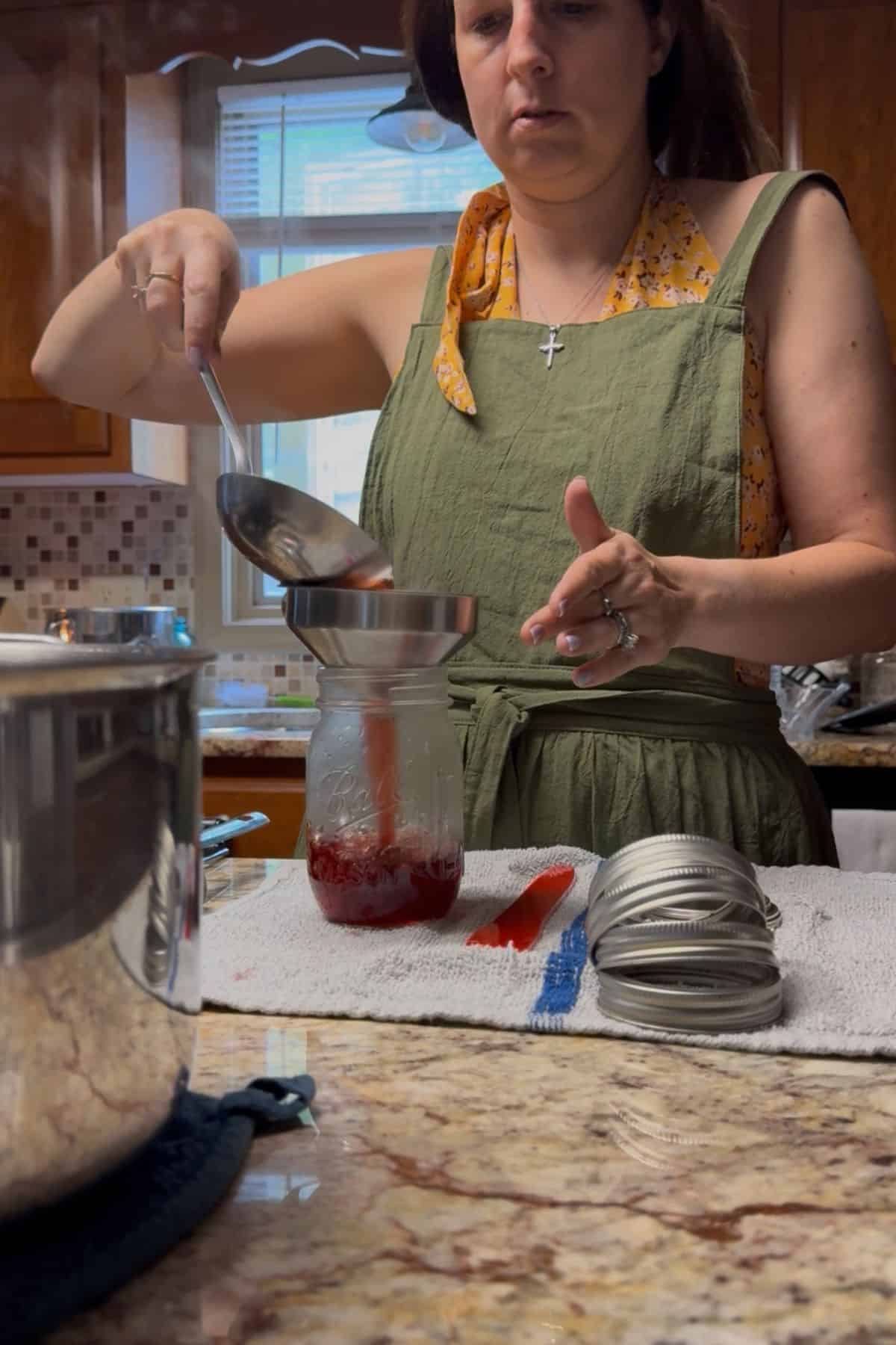 Woman ladling hot strawberry syrup into a mason jar using a funnel, preparing for water bath canning on a kitchen counter