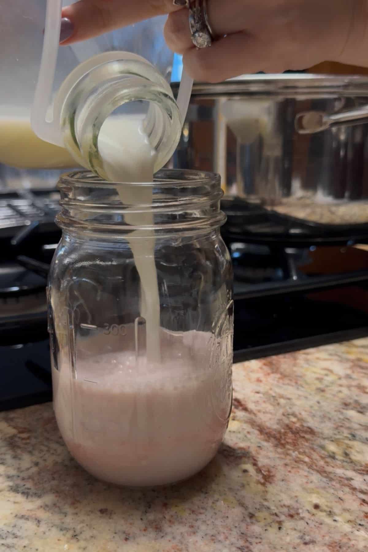 Milk being poured from a glass jug into a mason jar of strawberry syrup to make homemade strawberry milk on a kitchen counter