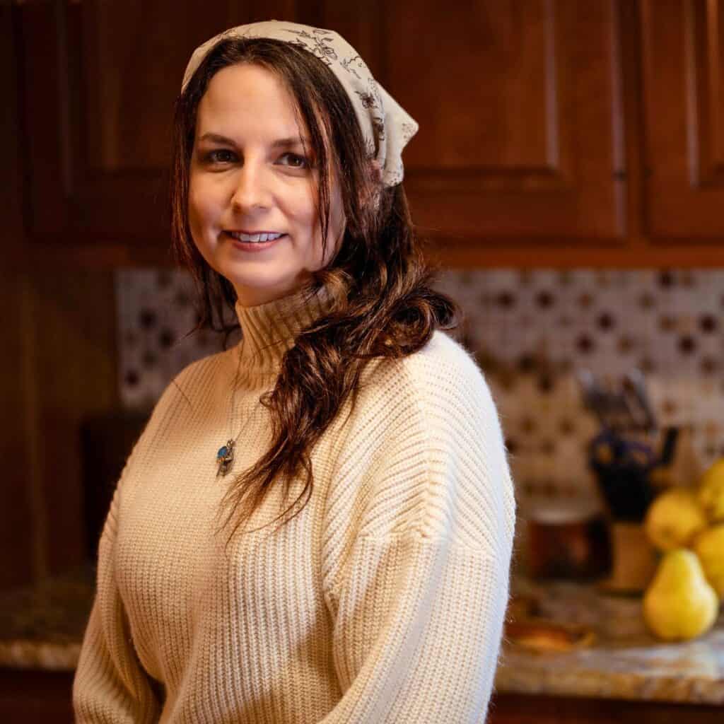 Elle, founder of The Rustic Elk, smiling in her warm farmhouse kitchen wearing a cream sweater and floral headscarf, representing faithful homemaking, simple living, and Catholic life.