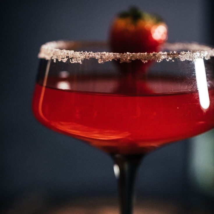 Close-up of a strawberry lemon drop martini in a coupe glass with a sugared rim and fresh strawberry garnish, photographed against a dark moody background.