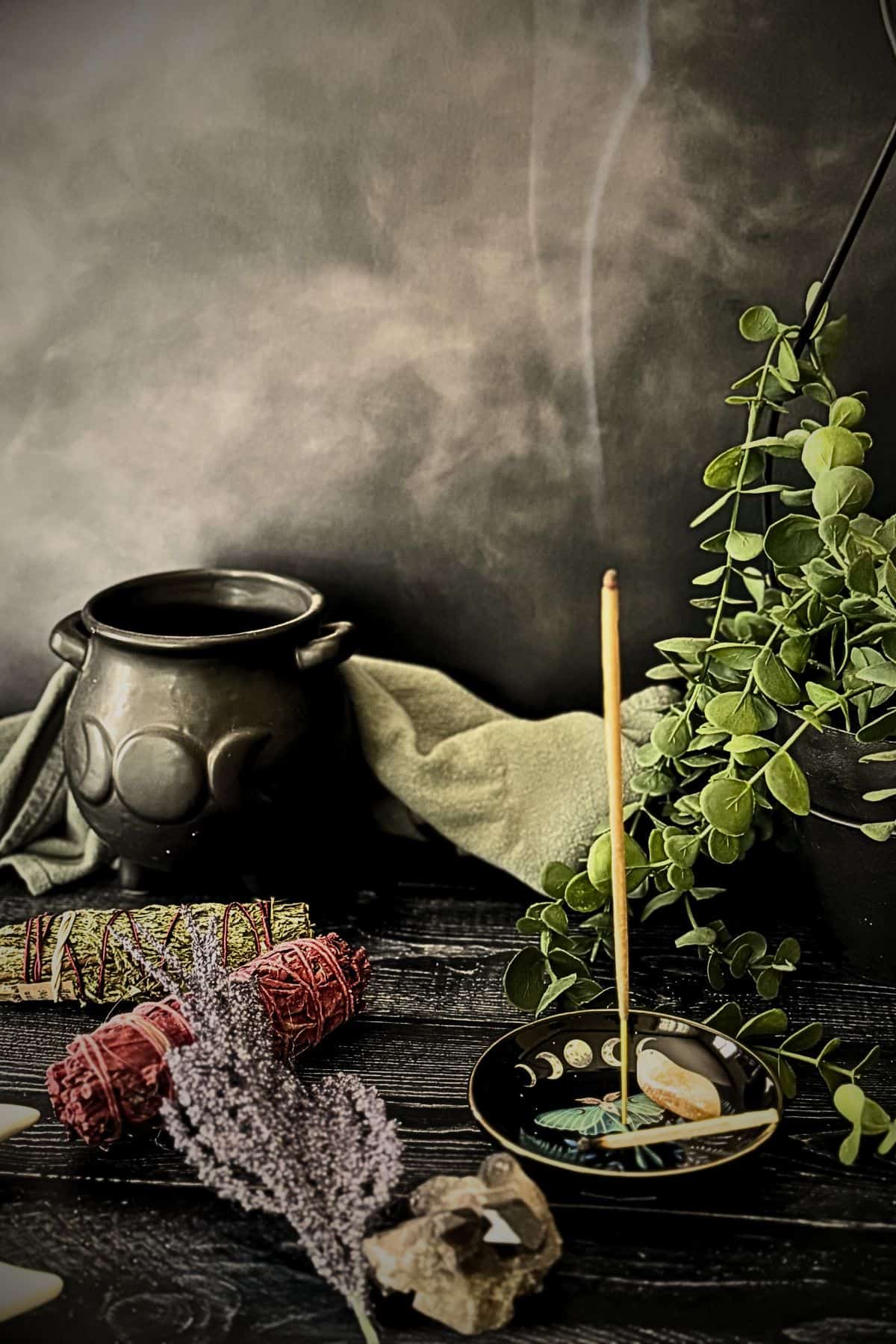 Smoking incense beside a black cauldron, crystals, herbs, and moon phase dish arranged on a dark wooden table.