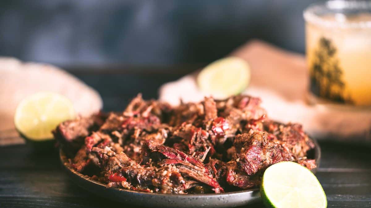 Smoked barbacoa shredded beef on a black platter with lime wedges, corn tortillas, and chipotle braising juices