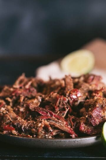 Smoked barbacoa shredded beef on a black platter with lime wedges, corn tortillas, and chipotle braising juices