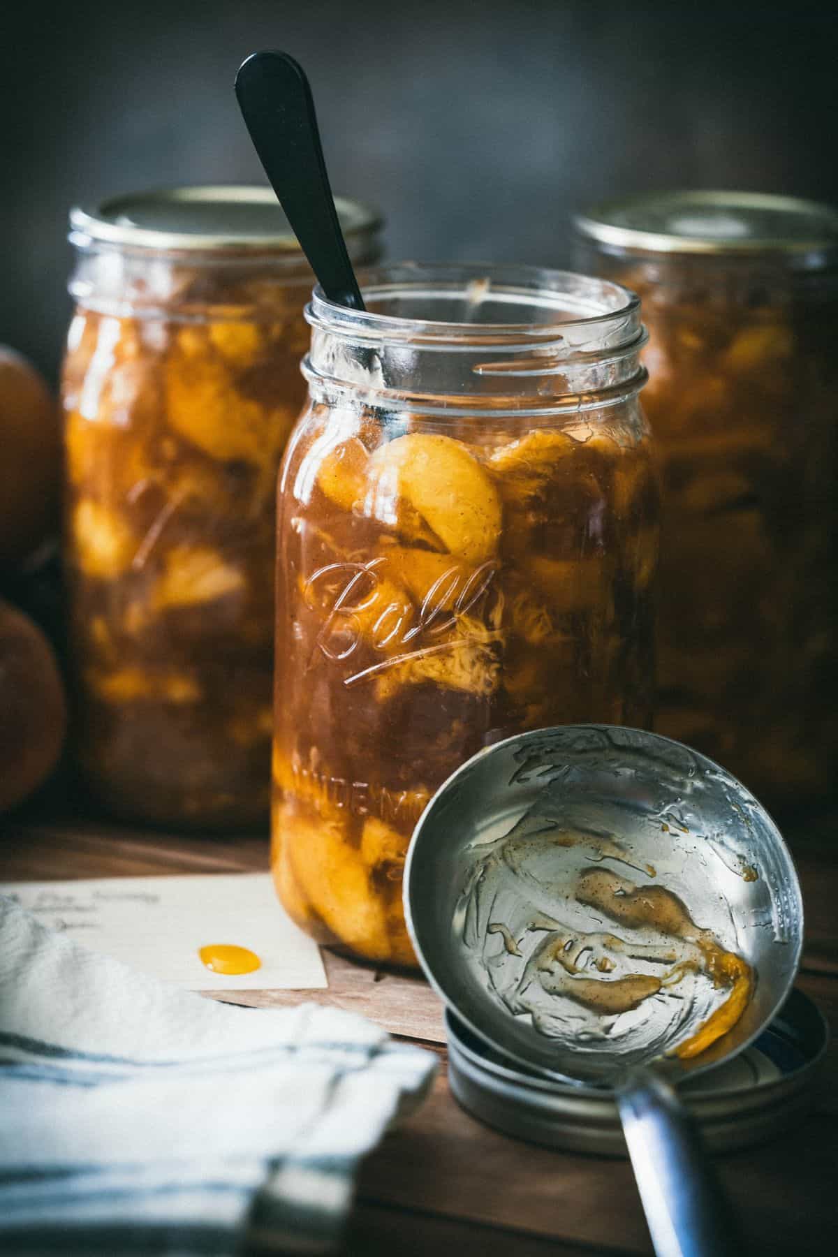 Open jar of homemade canned peach pie filling with cinnamon-spiced peaches in thick Clear Jel syrup beside sealed mason jars on a wooden board.